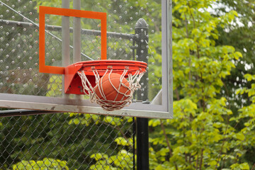 close up of a basketball going through the net of the basketball hoop with glass backboard visible and trees in background, a score in basketball game