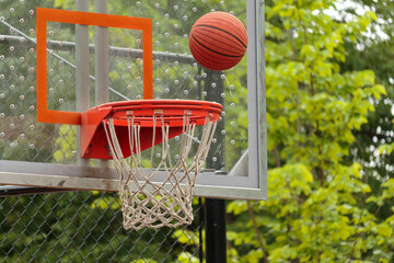 close up of a basketball hovering over the basketball hoop as it flies through the air with glass backboard visible and trees in background, suspended in air