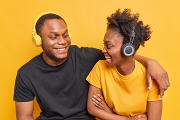 Obraz premium Joyful African American woman and man embrace and look with happy expressions at each other have fun listen music via headphones isolated over vivid yellow background. Friendship entertainment