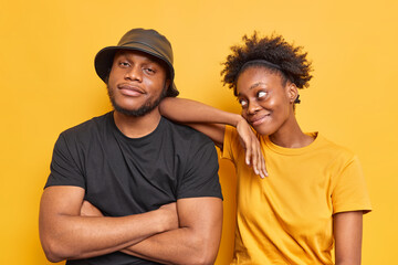 Fototapeta premium Indoor shot of woman and man best friends enjoy spending free time together dressed casually pose against yellowbackground. Pleased dark skinned curly female model leans at shoulder of boyfriend