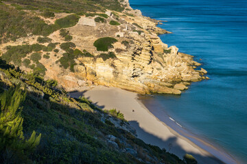 Obraz premium Single Person sitting on the beach with rock formation in golden sunlight from above with blue sea, Figueira, Portugal
