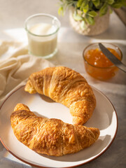 In the foreground of the photo, we see two croissants on a porcelain white plate. In the background, orange jam and milk in a glass glass. White silk tablecloth. Bright hues. Close-up.