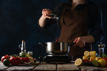 In the photo, the chef is preparing broth. There is a saucepan on a wooden table, in which the chef adds spices. The table also contains the ingredients used to make the broth.