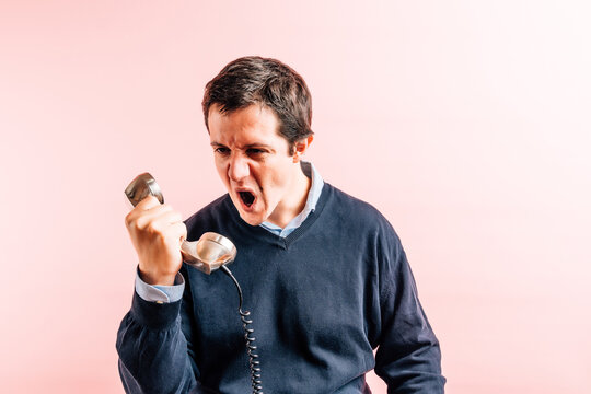 Young Adult Thirty Five Year Old Man In Blue V-neck Shirt And Sweater With Pink Background Angry Yelling At A Wired Phone