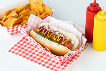 Close up of a hotdog in a red and white checkered tray with a bottle of ketchup and mustard and potato chips in behind. - obrazy, fototapety, plakaty