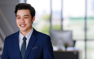 Naklejka na ściany i meble young attractive asian business man wearing navy blue suit with white shirt and necktie smiling beautifully looking in the future in office with natural lighting