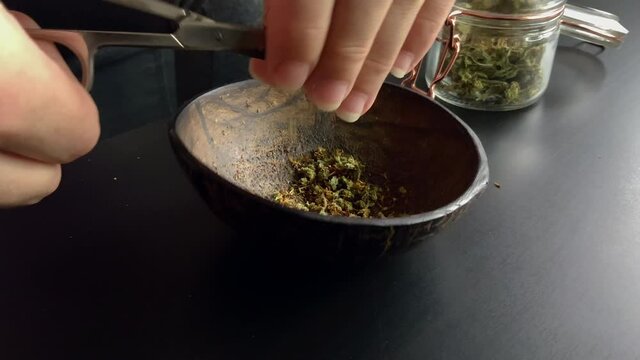 Close up of hands cutting weed in half coconut bowl, besides a jar full of weed.