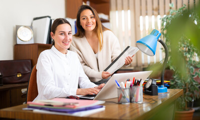 Confident woman assistant standing with documents near her female boss in office