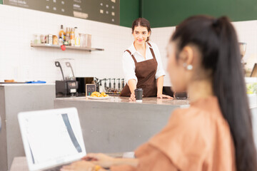 Beautiful caucasian barista woman places coffee cup on table of coffee bar for customer while other Asian customer woman using laptop