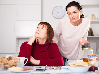 Two women having serious quarrel and talking at table in home interior