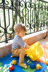 Little girl sits on a rug on the balcony and fumbles in a box with toys