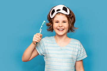 Happy child kid girl with sleeping mask brushing her teeth with toothbrush in morning isolated over blue background. Health care, dental hygiene. Mockup, copy space