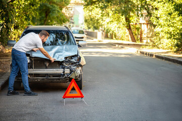 Man driver looking on Smashed broken car in accident. Red emergency stop triangle sign afore Destroyed car in car crash traffic accident on city road. Copy space