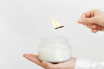 Womans hand uses match to light aromatic candle in glass jar with natural ingredients on white...