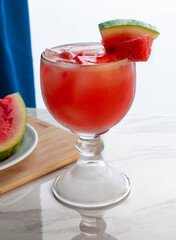 vertical watermelon cold drink with pieces of watermelon on top and in the background over a cutting board, side view
