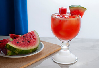 side view horizontal glass cup of watermelon cold beverage with slices of watermelon on the side on a white plate and over a cutting board, lit by a window on the background.
