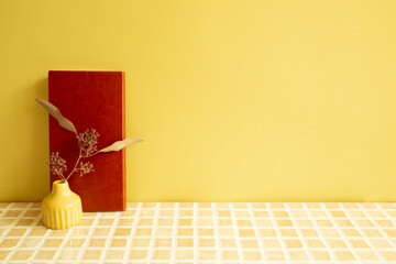 Notebook and vase of dry flowers on beige ceramic mosaic tile desk. yellow wall background. Work...