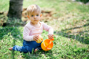 Little girl sits on a green lawn and holds a yellow bucket and an apple in her hands