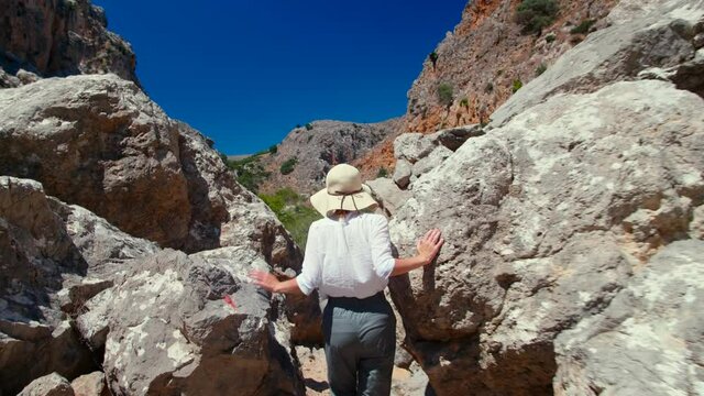 Traveler Female is Hiking Between Rocks in Zakros Gorge on Crete, Greece. Tourist Woman in Hat Explores Natural Mountains at Trekking on Vacation in Summer. 4K Hand Held Tracking Wide Shot