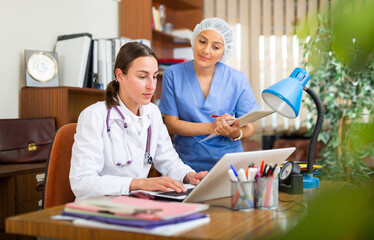 Confident woman doctor with assistant working in medical office using laptop computer, consulting patient online, telemedicine concept