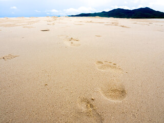 Beach sand footprints with copy space. Close up human footprint from walking barefoot on the sand beach heading to the mountain and blue sky background. Journey, hope, summer background concept.