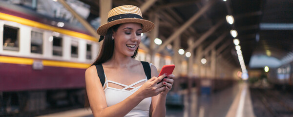 Young brunette woman traveler laughs while playing a mobile phone in train station platform. 20s...
