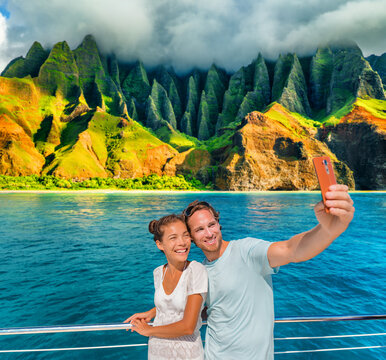 Hawaii cruise ship couple tourists taking selfie photo with phone at Na Pali Coast, Kauai island, USA summer vacation travel. Happy Asian woman, Caucasian man, interracial friends group of people.