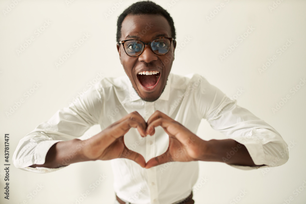 Wall mural Studio image of happy excited young dark skinned guy wearing spectacles opening mouth widely holding hands in front of him, making heart shaped gesture, being full of love, expressing affection