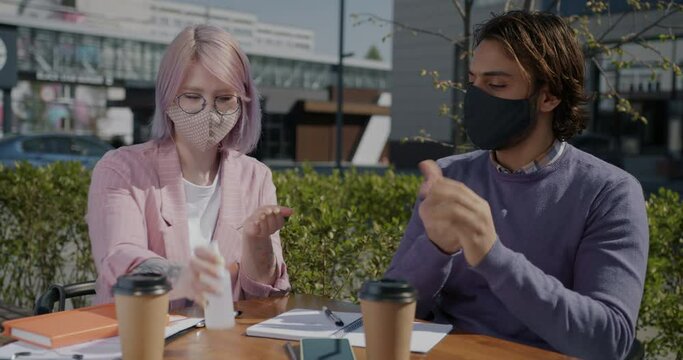 Students wearing masks are studying in street cafe spraying hands with sanitizer talking and laughing. People and corona virus pandemic concept.