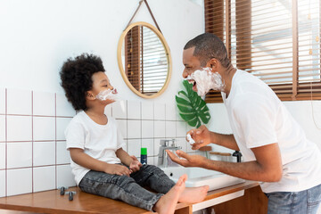 Funny Black African American Father and little boy laughing while playing shaving foam on their face in bathroom at home on holiday. Happy African family having fun