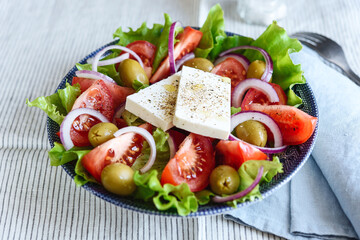Greek salad of fresh vegetable with tomatoes, lettuce, olives, red onion and feta cheese in bowl on white striped linen tablecloth. Selective focus