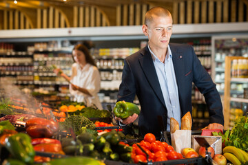 Young confident man carefully selects fresh vegetables on the counter in the supermarket