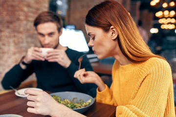 woman having dinner at a table in a cafe and a man with a cup of coffee in the background