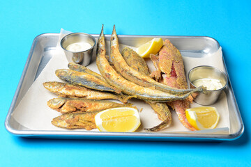 Fried fish with fresh herbs and lemon served on a metal tray over blue background.