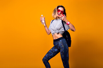 Slim girl with short hair with backpack going to gym and holding bottle of water. Laughing sporty young woman in sunglasses posing on bright background in studio with apple in hand.
