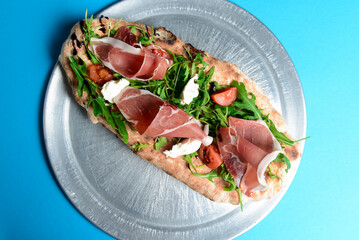 Sandwich with prosciutto, cheese and fresh arugula served on a metal tray over blue background.