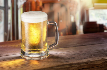 Cooled mug of pale beer with condensation drops on glass surface on the wooden table. Blurred bar at the background.