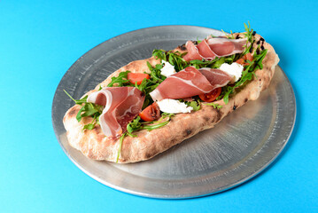 Sandwich with prosciutto, cheese and fresh arugula served on a metal tray over blue background.