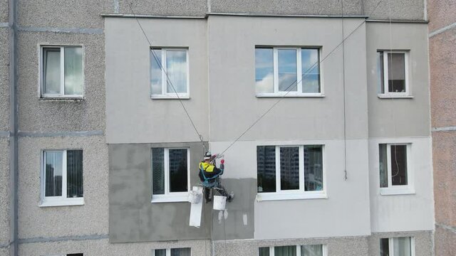 An industrial climber suspended on ropes and paints the wall of a building with a roller. A male worker performs work on the insulation of the facade of a multi-storey building. View from the drone.