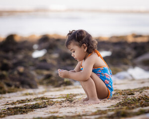 Cute little girl sitting on sandy beach, playing with sand. Happy childhood. Summer vacation. Holiday at the sea. Baby girl wearing blue swimsuit. Bali, Indonesia