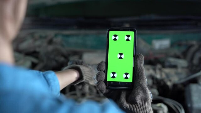 A garage worker holds a phone with a green screen in front of the open hood of the car. Repair of equipment and engine in a car workshop.