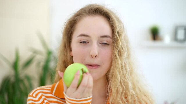Woman eat apple with playful smile, stand in living room home Spbas. Pretty blonde female bite