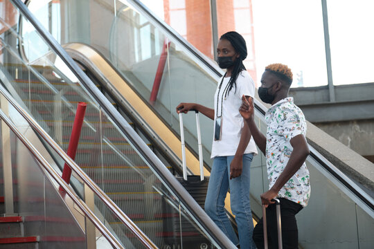 Young african couple wearing protective face masks and carrying suitcases climbing the escalator at the train station. - Powered by Adobe