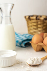 Close-up of wooden spoon with flour, milk bottle, eggs, basket and cloth, on white table, selective focus, white background, vertical