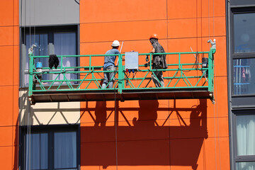 Worker standing on lifting platform near the wall of high-rise house. Construction works,...