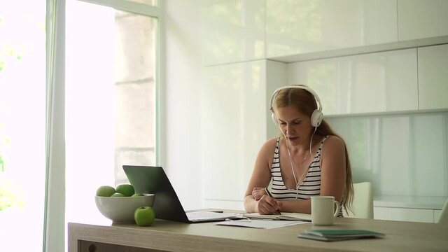 Woman in headphones works in pen in hands in front of laptop in white interior Spbd.