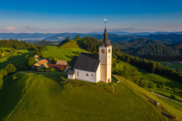 Fototapeta na wymiar Sveti Andrej, Slovenia - Aerial drone view of Saint Andrew church (Sv. Andrej) at sunset in Skofja Loka area. Summer time in the Slovenian alps with clear blue sky