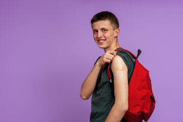 Happy Smiling Vaccinated Young Man Student, With Backpack Showing His Hand With Patch After Injection Covid-19 Vaccine, Poses On Purple Background. Advertising of vaccination against coronavirus.