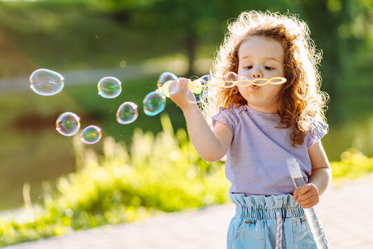 Little curly girl blowing soap bubbles at warm summer day in the park