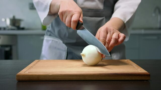 Female hands cut white onion bulb into halves by knife on wooden board